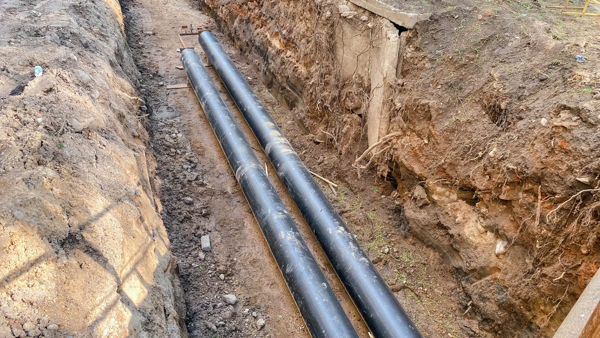 Two black plastic pipes laid along the floor of a deep trench beside a building